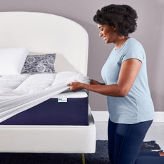 Young woman kneeling next to a bed and pulling the quilted fiberfill stretch cover over a mattress corner 